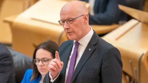 John Swinney is wearing a dark suit and purple tie. He points his finger diagonally up while standing to address the Scottish parliament 