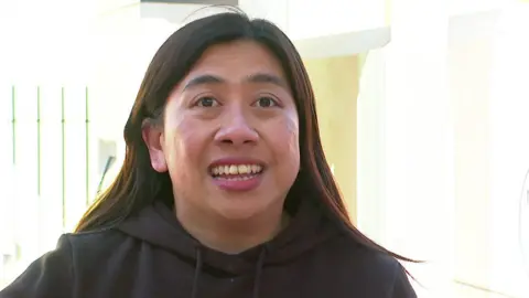 A young woman with long brown hair smiles while wearing a brown hoodie.