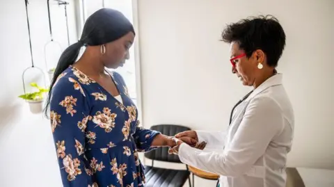 Getty Images A doctor examines a woman's hand and wrist in a consulting room. The doctor has a white coat and is wearing glasses and the patient is wearing a blue dress and has her hair in a pony tail. The walls are light and the room is in bright daylight, with plants hanging in pots form a rail.