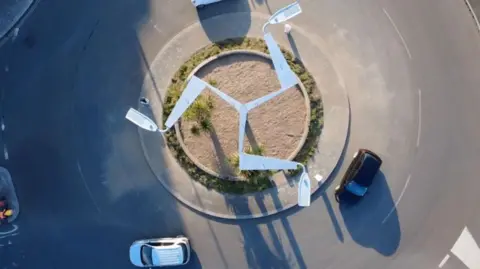 An aerial shot of a silver lighting column is silver has a three legs sculpture on top. It stands in the middle of a roundabout with cars circling.
