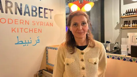 Mouna Elkekhia sees at the camera. Above her are two lights and behind her a kitchen and a sign which reads "Arnabeet, Modern Syrian Kitchen" in English and Arabic