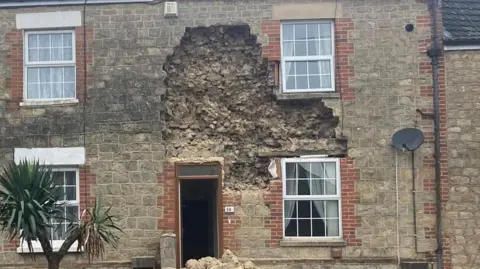 A terraced house which has partially collapsed. A large amount of bricks can be seen missing from the front. The house is cordoned off by a yellow gate. A pile of bricks can be seen by the front door. 