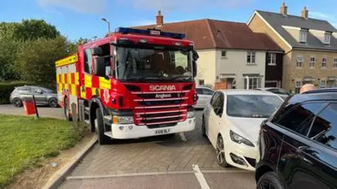 Essex County Fire and Rescue Service A fire engine is trying to turn around a corner, but has had to go over a grass verge due to the parking of a car on the other side of the road.