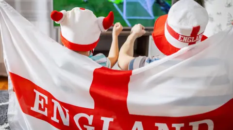 Getty Images Two young children wrapped in an England flag and wearing red and white hats watching a football match on the TV.