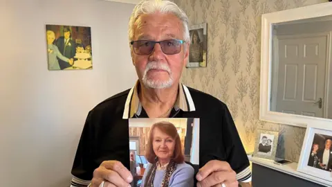 A man in his 70s is stood in the middle of a living room holding up a picture of his deceased wife. The man has grey hair and a beard and is wearing glasses.  The picture of his wife shows a lady in her 70s with shoulder-length brown hair.  There are pictures of the couple on the wall and on the mantelpiece.