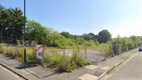 Google An area of land that is fenced off with metal fencing. There is a "sold" sign on the fence. Beyond the fence are overgrown plants, grass and weeds, as well as piles of gravel and dirt.