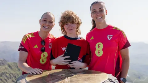 FAW Three women stood at the top of Yr Wyddfa mountain. They are all wearing red Wales shirt and smiling at the camera. One is blonde, one has brown hair, and the one in the middle ahs curly red hair and is holding a black notebook.