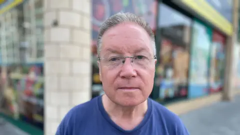 Laura Foster/BBC Mark is wearing glasses. He is standing in Basildon Town Centre