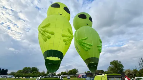 Fly Alien Two alien hot air balloons tethered to vehicles on a field.