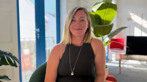Charlie Cox - A person with shoulder-length light hair sits facing the camera in a modern, sunlit room. The person is wearing a sleeveless dark ribbed top and a long pendant necklace while seated in a green chair. Large green houseplants frame the scene, and blue-framed glass doors are visible on the left. In the background, a television, a chair with a red cushion, and bright natural light create a warm indoor setting.