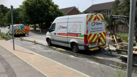 BBC Two white vans parked on the road, with two men in bright orange uniform walking along. 