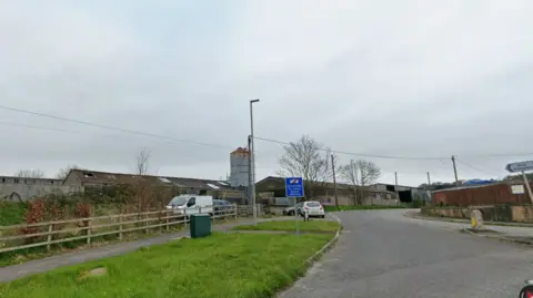 Google A google streetview of a road with a farm buildings on one side and industrial units behind a fence on the other
