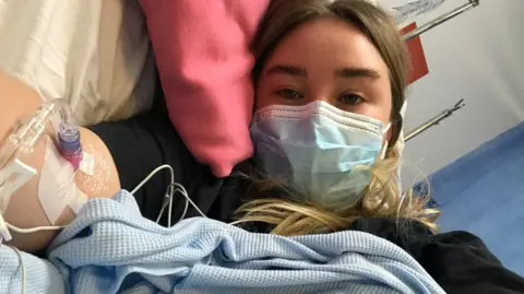 Chloe Dickson lies in a hospital bed with a drip connect to her inner upper arm. She lies on her side wearing a blue face mask covered in a blue blanket. She rests her head on a pink pillow and looks directly into the camera.