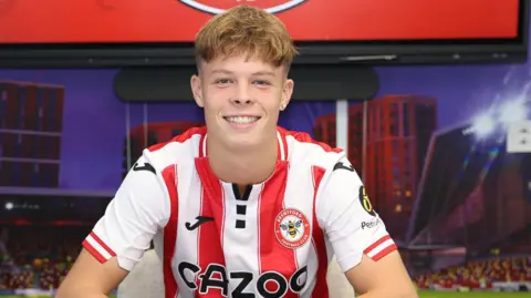 Pro Sports Images Luca is wearing a Brentford Football Club jersey with red and white vertical stripes sits at a table, signing a document. Another identical jersey is laid out on the table in front. The background shows a large screen and stadium graphics.