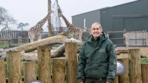 West Midlands Safari Park Lisa Watkins with blonde hair is smiling and wearing a green coat outside the giraffe section. Two giraffes look symmetrical in the background.