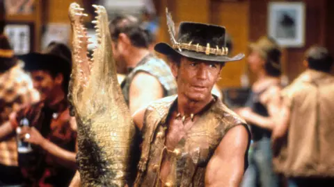 Getty Images Paul Hogan dengan buaya mati palsu saat syuting buaya Dundee. Dia mengenakan topi dan rompi.