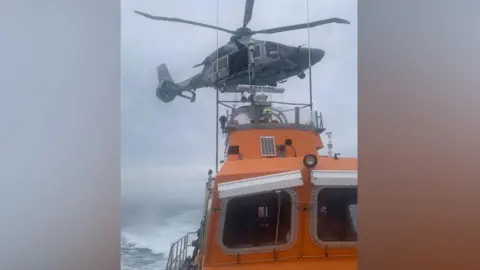 A grey helicopter hovers over an orange lifeboat in the middle of the sea during a rescue operation. It is a cloudy day. A man wearing a yellow helmet is on the helm of the boat.