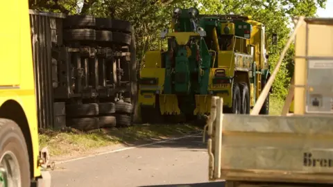 The underside of a lorry can be seen as the vehicle is on its side on the side of the road. A tow truck is being lined up. 