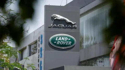 A green sign on a large grey block which reads Land Rover and a silver sign above it which says Jaguar. in the background there is a large grey building and there are leaves in the foregroun