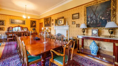 Victor Mee Auctions The dining room in the Bishop's House in Newry.  It is an ornate wallpapered room with several framed paintings, a marble fireplace, a long wooden dining table and other furniture and ornaments. 