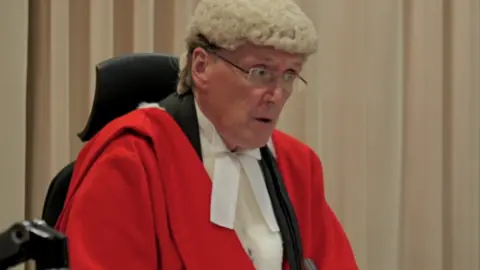 A seated man wearing traditional judicial attire including a bright red robe over a white shirt and white neck bands, along with a curled, light-coloured ceremonial wig. 