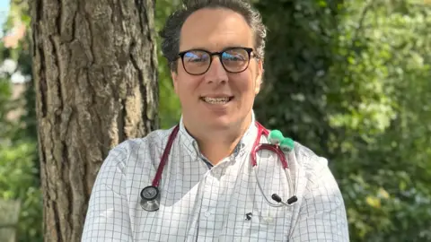 Health Plus The image shows Dr Ed Klaber standing outdoors in front of a tree with greenery in the background. Dr Klaber is wearing a white and black checked shirt. He is wearing glasses and has a stethoscope around his neck.