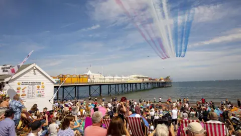 Crowds of people sat on Paignton beach admiring a Red Arrows display overhead, the blue sea in the distance alongside Paignton pier, on a sunny day.