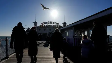 Reuters A pier with people in coats walking along it. The sun shines overhead and a seagull is flying over it.