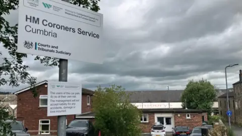 A sign for HM Coroner's Service Cumbria outside the brown brick building. There is a car park in front and there is a partially visible, orange sign for a Sainsbury's next door. 