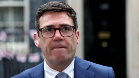 Close-up of Andy Burnham wearing tortoiseshell glasses, a light blue suit and tie and white shirt, standing in front of a blurred No 10 Downing Street