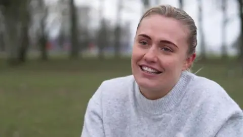 Gemma Richardson, a 23-year-old woman, sits in a park with a green field and trees behind her. She is smiling with blond hair, tied back, and is wearing a light grey sweater.