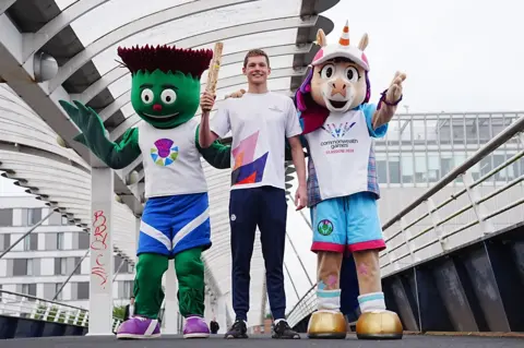 PA Media Two mascots standing either side of a young man holding a wooden baton on a bridge. The mascots are a thistle and a unicorn, both with smiling faces and wearing Team Scotland uniforms.