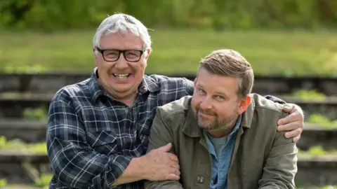Graeme Hunter / Happy Tramp North Gregor Fisher and Greg McHugh smiling at the camera. Fisher has his arm around McHugh. He wears a blue and white plaid shirt and glasses, while McHigh is wearing a green jacket over the top of a blue shirt.