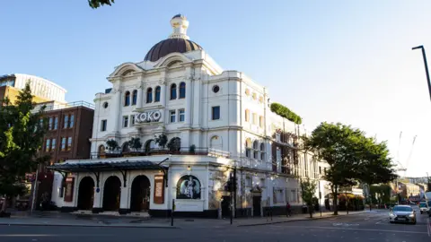 Koko/PA Media Wide shot of the outside of Koko music venue, a white building formerly known as the Camden Palace that sits on the corner of the road. The facade is in a Baroque pastiche style with three arched entrances and an awning above them.