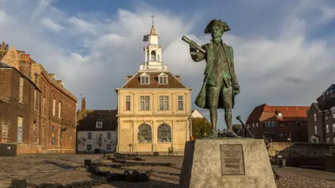 A statue of Captain Vancouver stands on a plinth. The bronze statue is wearing a tricorn and clutching a scroll. Behind him is the Custom House, a 17th century three storey building, topped by a white tower.