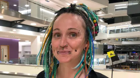 Jon Wright/BBC Jessie Carter smiles at the camera inside a council building. She has different coloured dreadlocks with multi-coloured beads in them. She has several nose, lip and facial piercings. 