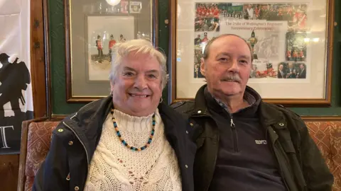 A silver-haired lady wearing a blue coat and cream, cable-knit jumper sitting next to a man with a silver-grey moustache and wearing a black jacket and dark, navy blue fleece. 