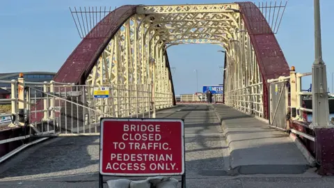 A red road sign that reads Bridge closed to traffic, Pedestrian access only. IT is placed on the road in front of the roadway onto the swing bridge, which is a red and white metal structure.