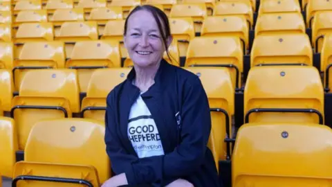 Wolves Foundation A woman with dark hair tied in a ponytail sits in a football stand that has gold-coloured seats. She is smiling and wearing a white T-shirt that reads "Good Shepherd", with a navy jacket with the same name on it.