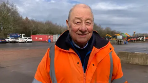 A man in an orange fluorescent jacket smiles in a large carpark with storage containers and other vehicles in the background.