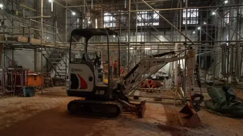 Inside a building, filled with scaffolding. You can see an excavator and people working in hard hats in the background.