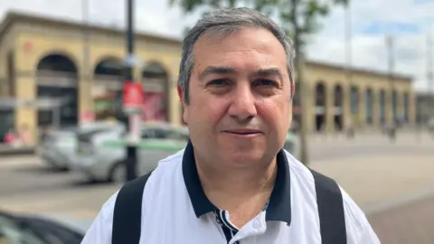 Cenk Sözen looking at the camera with a slight smile. He is wearing a white polo shirt. Behind him is the railway station building and taxi rank, out of focus. 