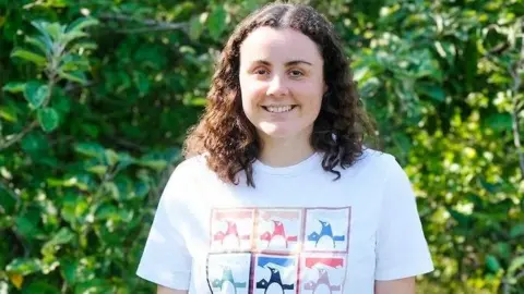 Maggie Coll - smiling and wearing a white t-shirt with a penguin graphic and the words 'Port Lockroy Antarctica'