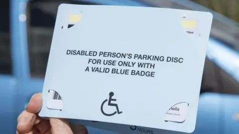 A hand holding a blue badge. On the front of the badge it says, DISABLED PERSON'S PARKING DISC FOR USE ONLY WITH A VALID BLUE BADGE. In the background of the picture is a blue car.