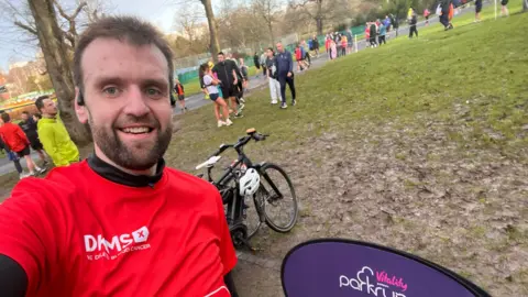 A man with dark hair and a dark beard wears a red T-shirt and takes a selfie in a muddy park after completing a park run with other runners stood behind him.
