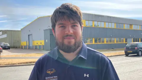 A man with a beard smiling at the camera. He is wearing a blue shirt and standing in front of a building with yellow lines to outline the windows