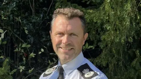 A man in white shirt and black tie with police badges on black epaulettes, smiling. He has black hair and is standing outside in front of a bush.