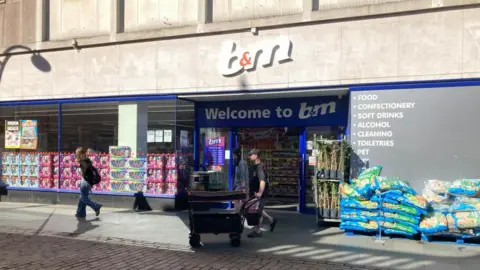Alex Pope/BBC The front of B&M, showing shoppers coming out of it. There are bags of items to the left. There is a white and orange sign above and plants also to the left. 