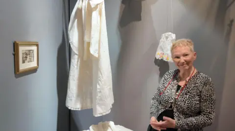 Myra Gilbert stands by a hanging collection of clothing, with a small frame on the left with an old family photograph on the wall. She is smiling and wearing a leopard-print patterned top. 