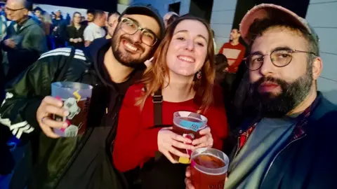 Two men and a woman hold plastic pints of beer at a crowded venue. The woman is wearing a red top and black dungarees. The men wear jackets, baseball caps, glasses and both have dark beards.